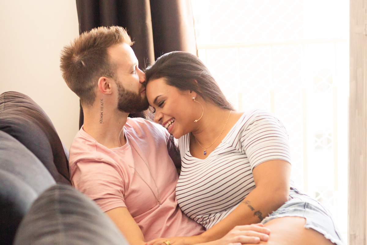 Foto do casal no sofá de casa sorrindo espontaneamente e fazendo carinho e beijando a testa, no ensaio lifestyle no bairro Vila Ema na cidade de SP