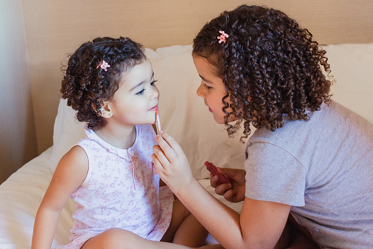 foto de irmãs se maquiando com pincel e blush e batom no quarto sentadas na cama no ensaio lifestyle em casa na cidade de São Paulo