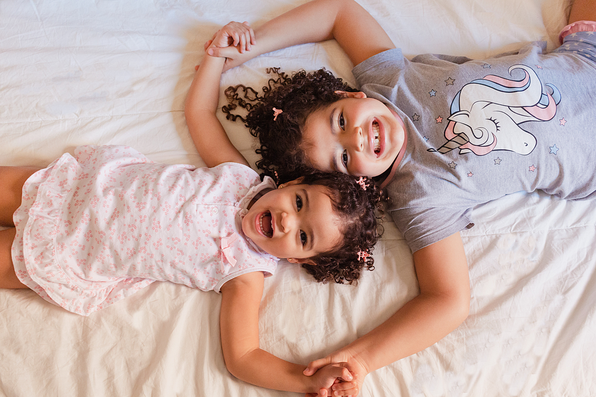 Irmãs sorrindo de mãos dadas na cama dentro do quarto no ensaio lifestyle infantil na Vila Ema cidade de São Paulo