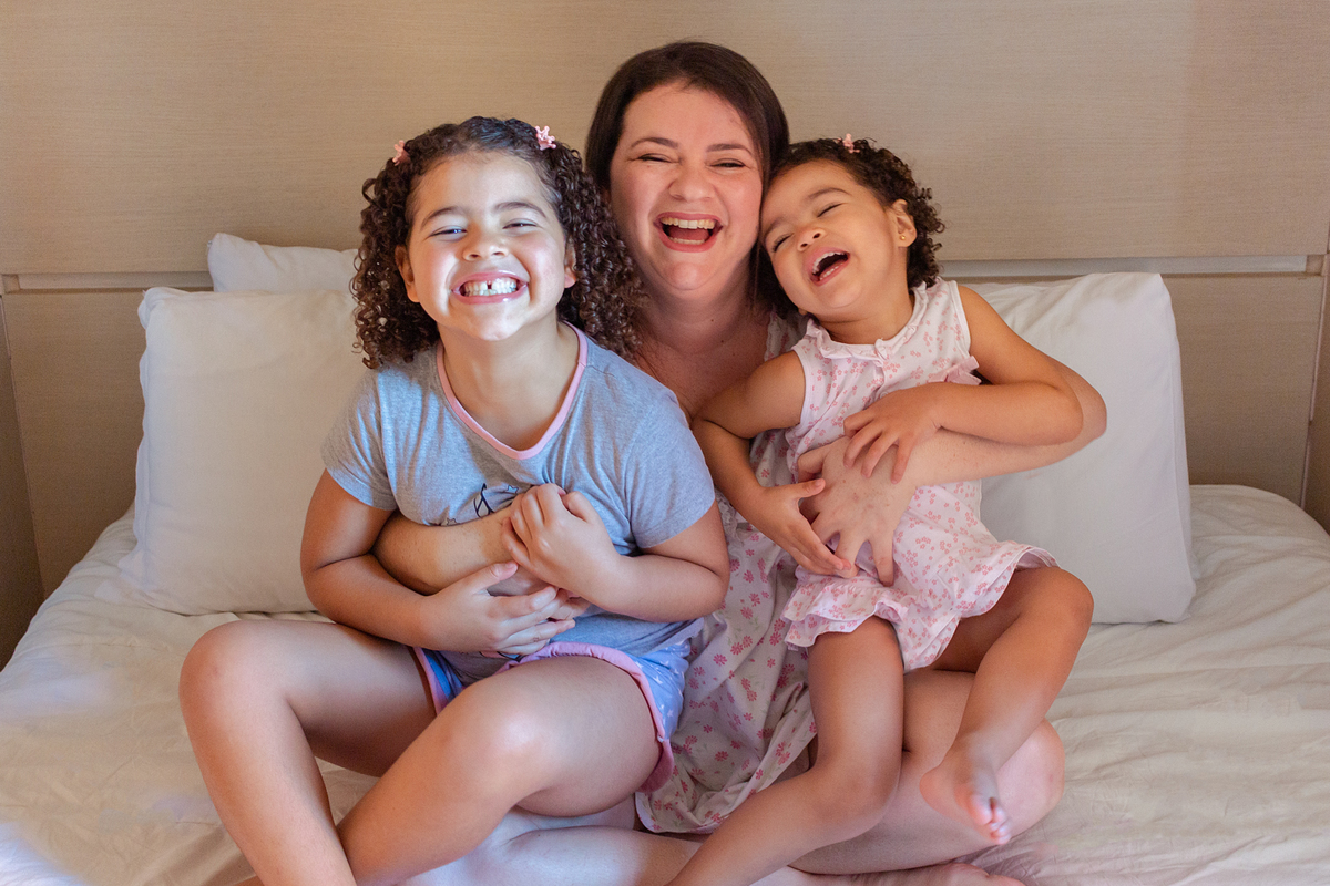 Mãe de menina abraçando as filhas na cama e sorrindo no ensaio de família em casa na Vila Ema zona leste de São Paulo
