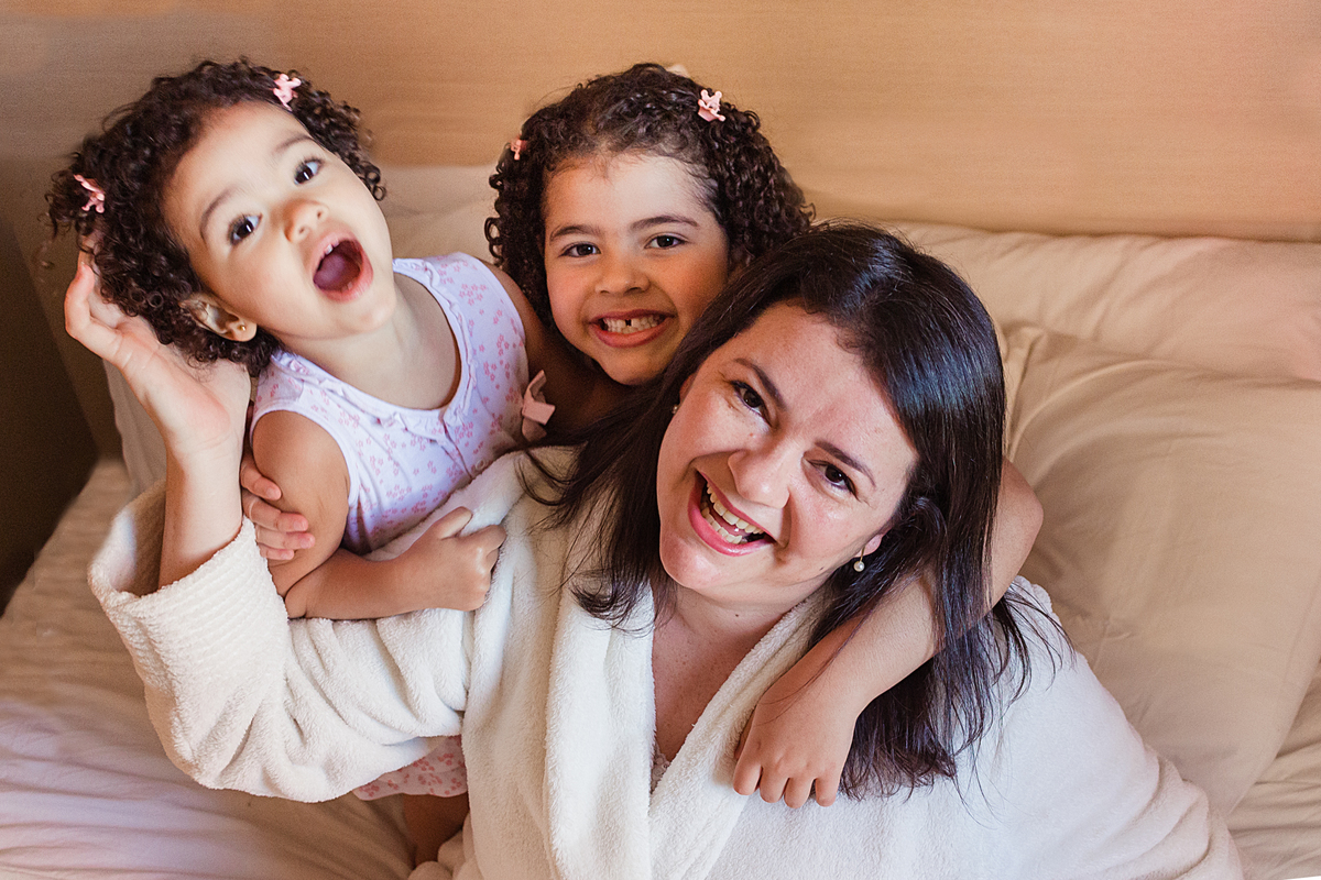 Mãe de menina abraçando as filhas na cama e sorrindo no ensaio de família em casa na Vila Ema zona leste de São Paulo