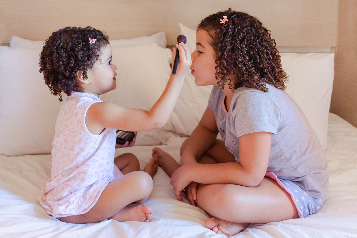 foto de irmãs se maquiando com pincel e blush no quarto sentadas na cama no ensaio lifestyle em casa na cidade de São Paulo