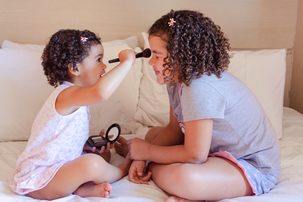foto de irmãs se maquiando com pincel e blush no quarto sentadas na cama no ensaio lifestyle em casa na cidade de São Paulo