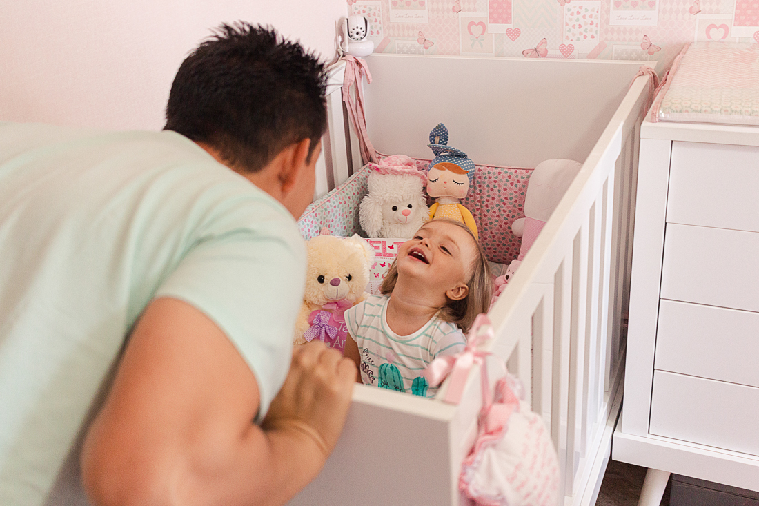 Pai brincando com a filha que está no berço  abraçando filha no quarto rosa de menina na cidade São Paulo