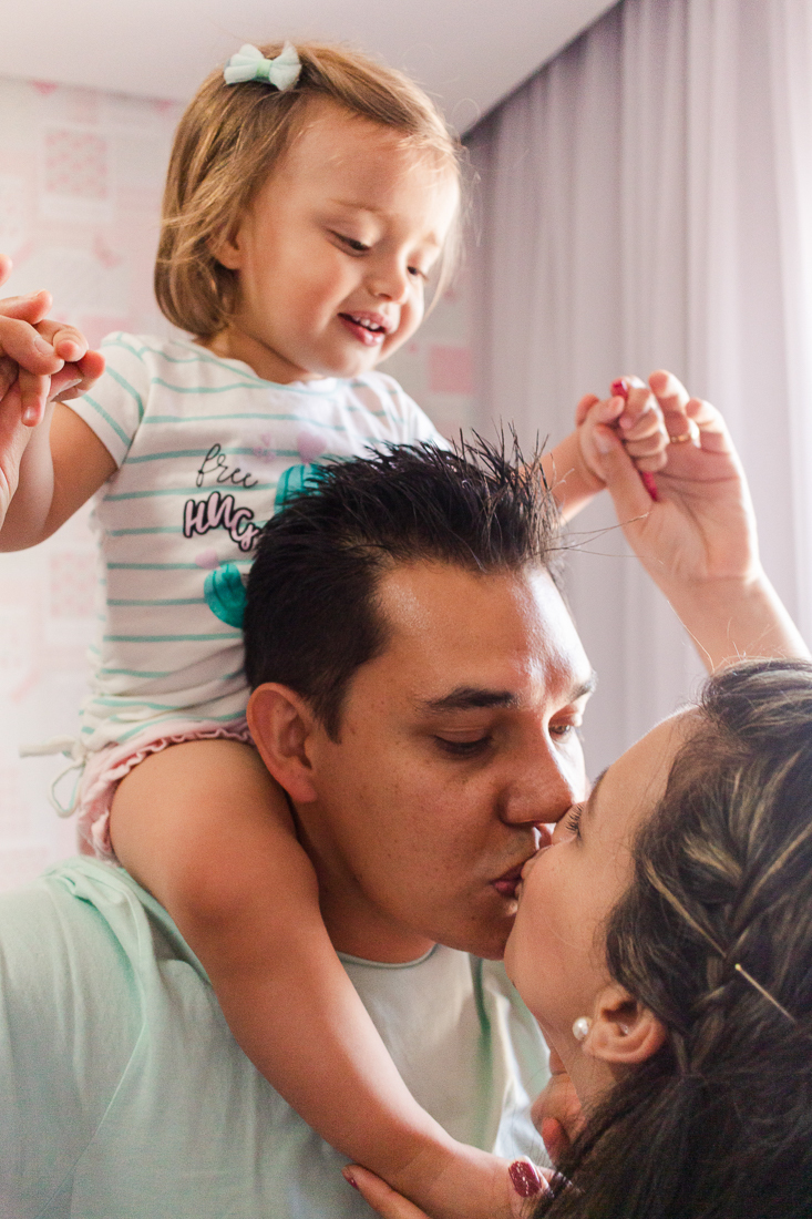 Pais se beijando com a filha no pescoço olhando, no ensaio de família na cidade de São Paulo