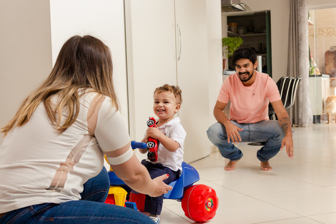Foto de pais brincando com a criança que está andando de motoca na sala de casa  no ensaio de família na cidade de São Paulo