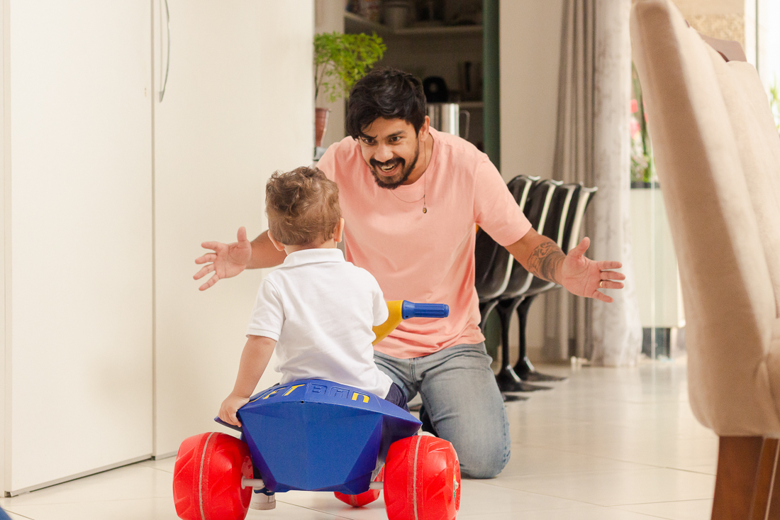 Foto de pais brincando com a criança que está andando de motoca na sala de casa  no ensaio de família na cidade de São Paulo