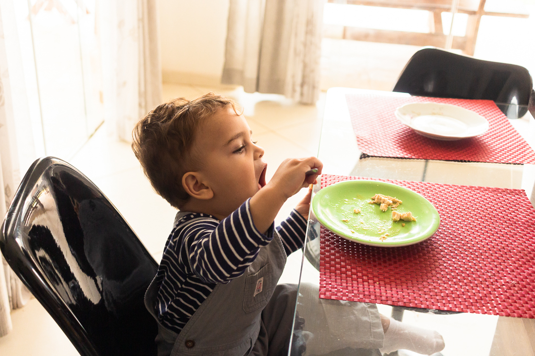 Menino Fernando comendo sozinho na mesa da cozinha no ensaio infantil em casa