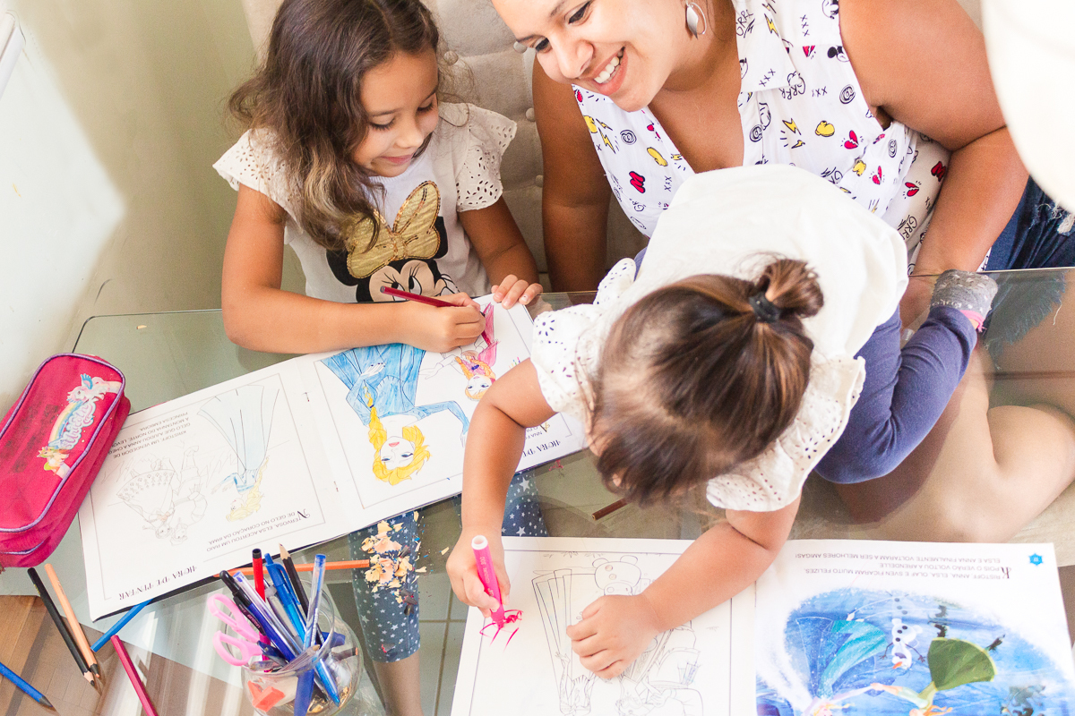 Foto de mãe e filhas fazendo lição e pintando o livro na mesa de casa no ensaio lifestyle infantil na cidade de Franco da Rocha em São Paulo