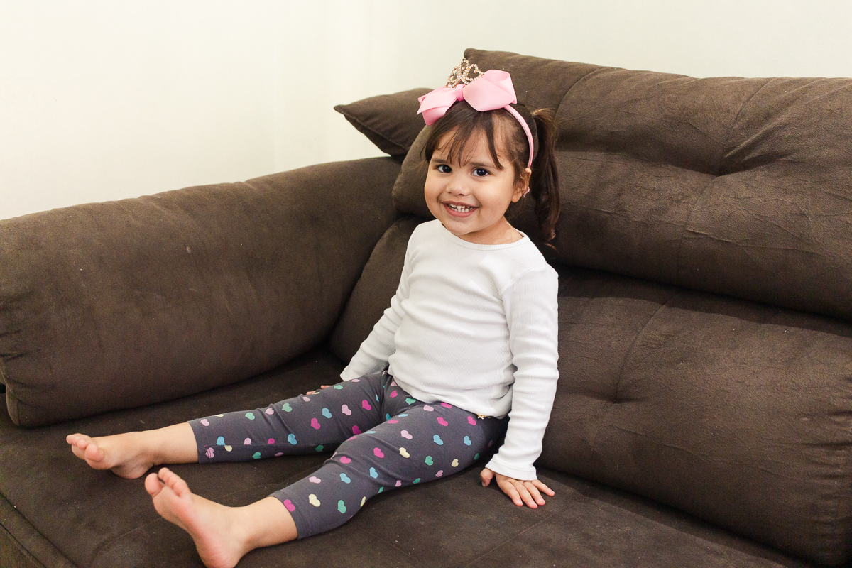 Foto de menina sorrindo sentada no sofá na sala de  casa no ensaio lifestyle infantil na cidade de Franco da Rocha em São Paulo