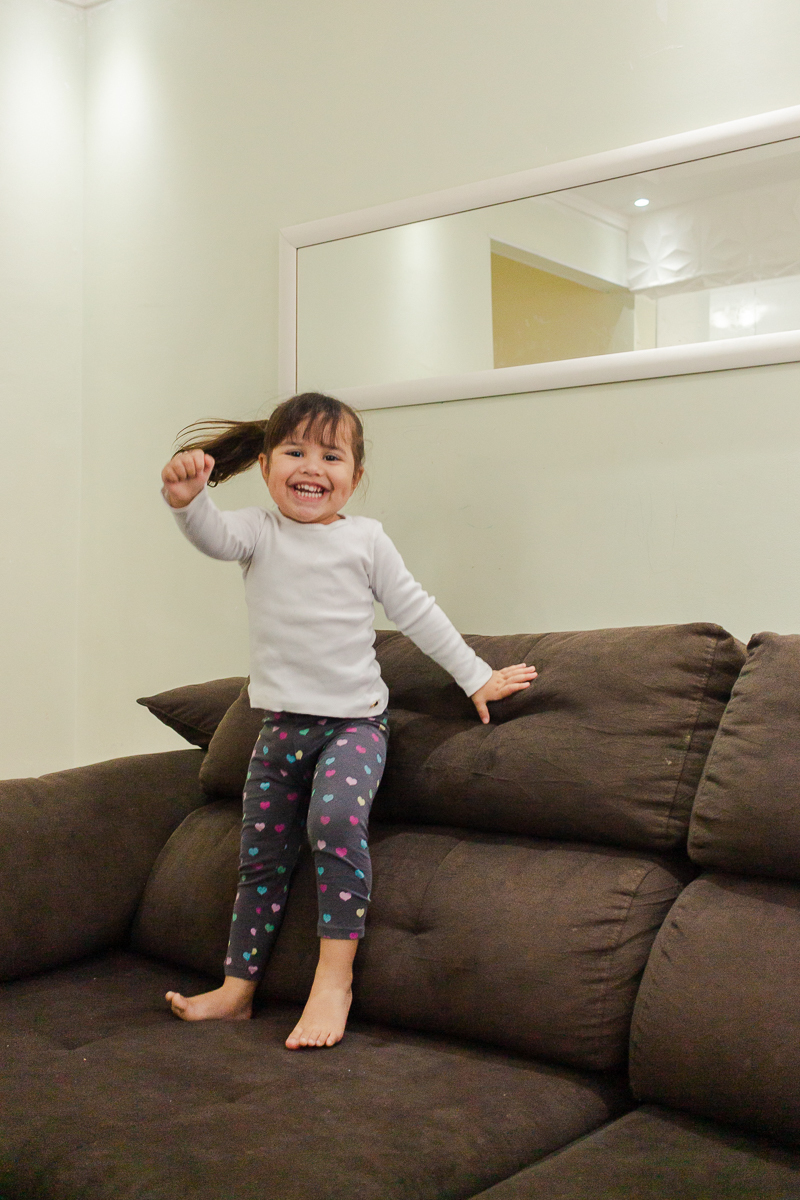 Foto de menina sorrindo brincando de pular sofá na sala de  casa no ensaio lifestyle infantil na cidade de Franco da Rocha em São Paulo
