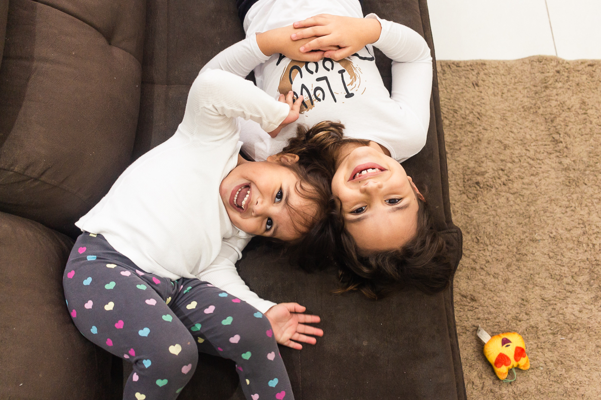 Foto de irmãs se olhando e sorrindo deitadas no sofá  da sala em casa no ensaio lifestyle infantil na cidade de Franco da Rocha em São Paulo