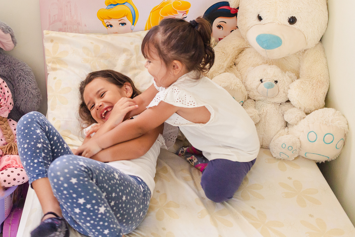 Foto de irmãs e mãe brincando de fazer cosquinhas na cama do quarto no ensaio infantil em casa na cidade de Franco da Rocha- São Paulo