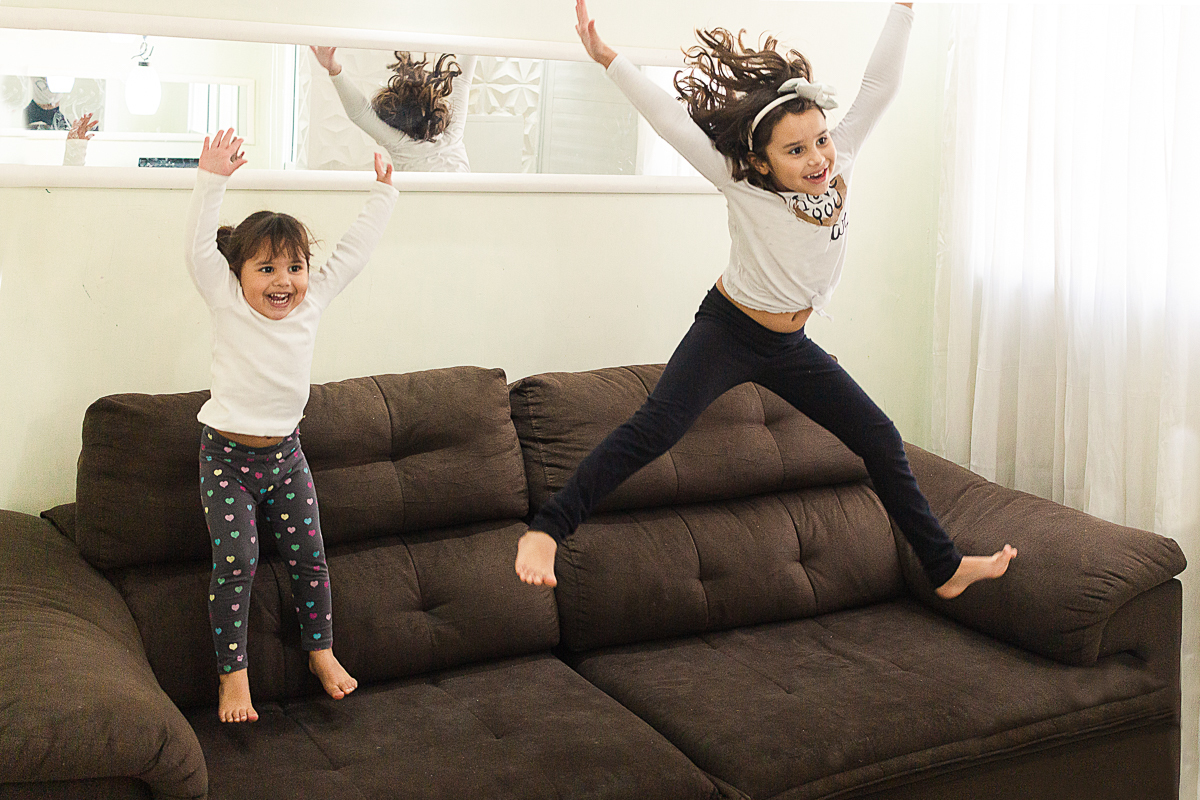 Foto de irmãs pulando e brincando no sofá de casa no ensaio lifestyle infantil na cidade de Franco da Rocha em São Paulo