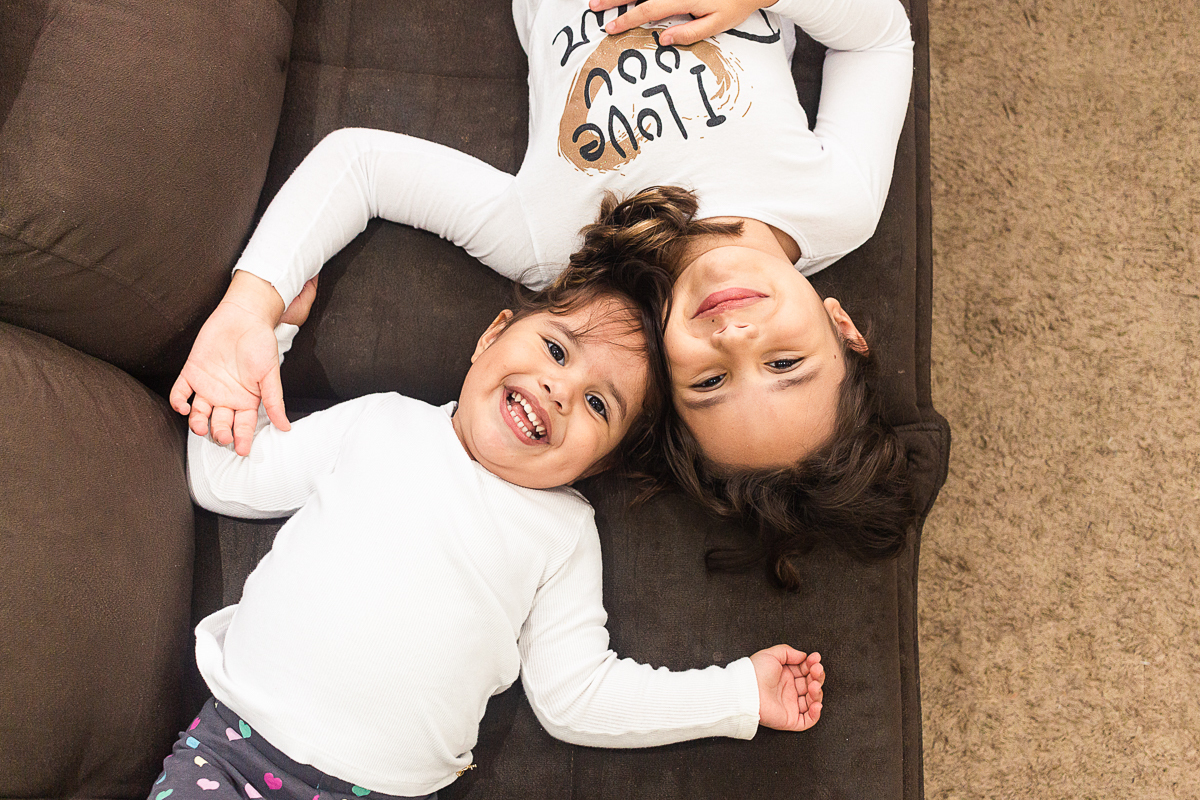 Foto de irmãs se olhando e sorrindo deitadas no sofá de casa no ensaio lifestyle infantil na cidade de Franco da Rocha em São Paulo