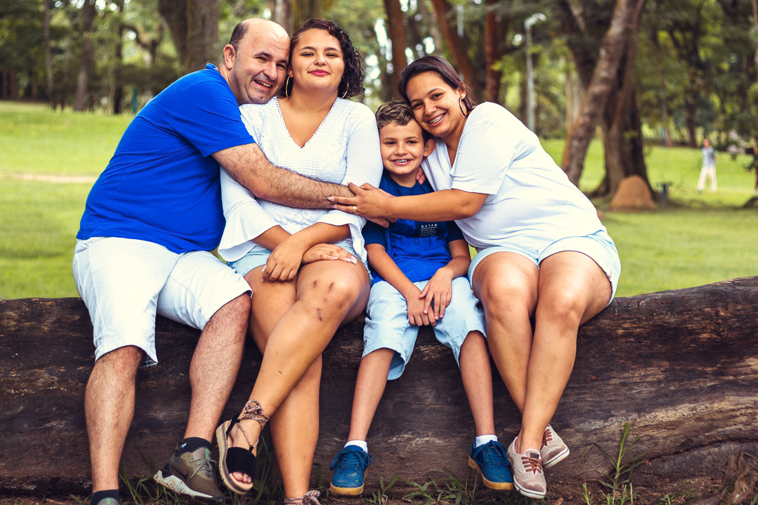 Foto de pais e filhos juntos sentados no tronco da árvore se abraçando de mãos dadas no ensaio externo lifestyle no parque do Carmo em São Paulo