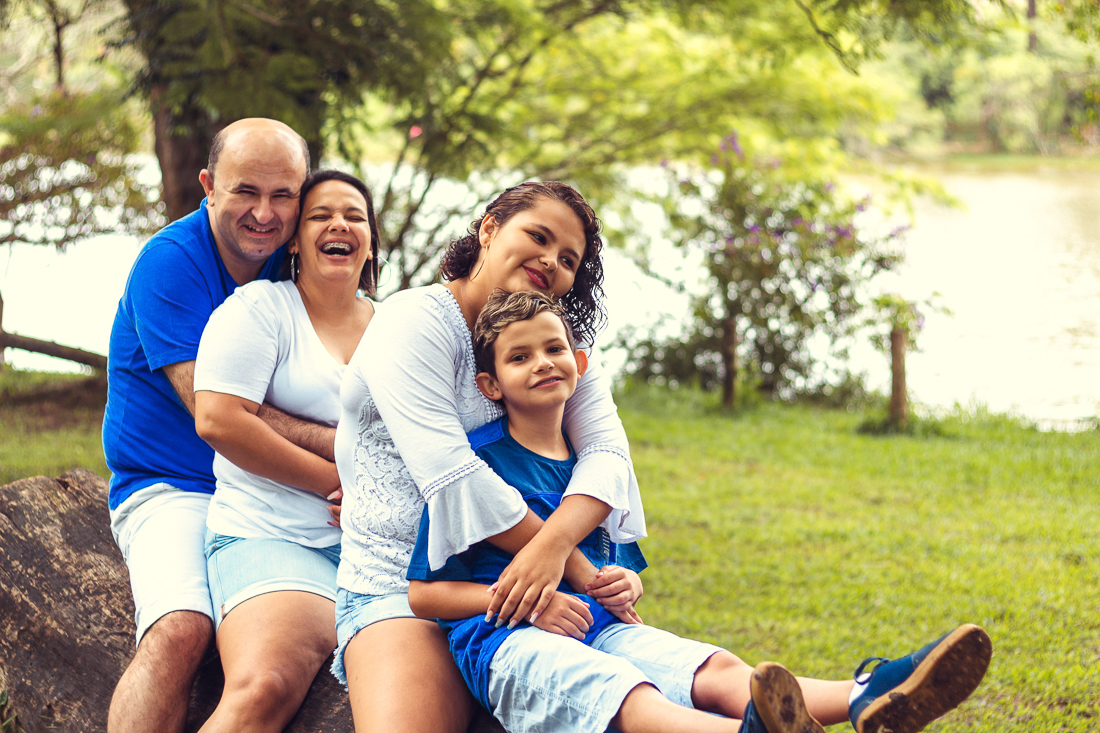 Foto de pais e filhos juntos sentados no tronco da árvore se abraçando de mãos dadas no ensaio externo lifestyle no parque do Carmo em São Paulo