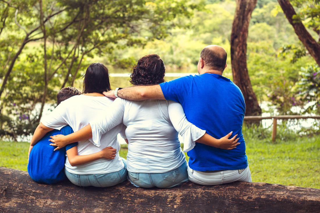 Foto de pais e filhos juntos sentados no tronco da árvore se abraçando de costas no ensaio externo lifestyle no parque do Carmo em São Paulo