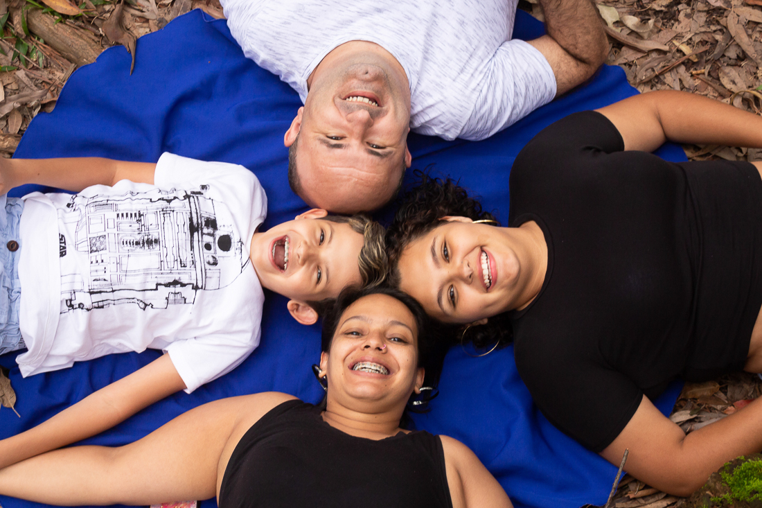Foto de pais e filhos deitados n grama com as cabeças próximas olhando para o alto no ensaio de família no parque do Carmo na Zona leste de São Paulo.