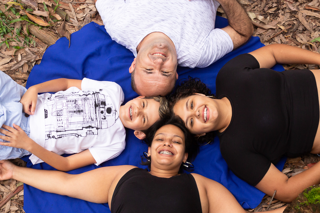 Foto de pais e filhos deitados n grama com as cabeças próximas olhando para o alto no ensaio de família no parque do Carmo na Zona leste de São Paulo.