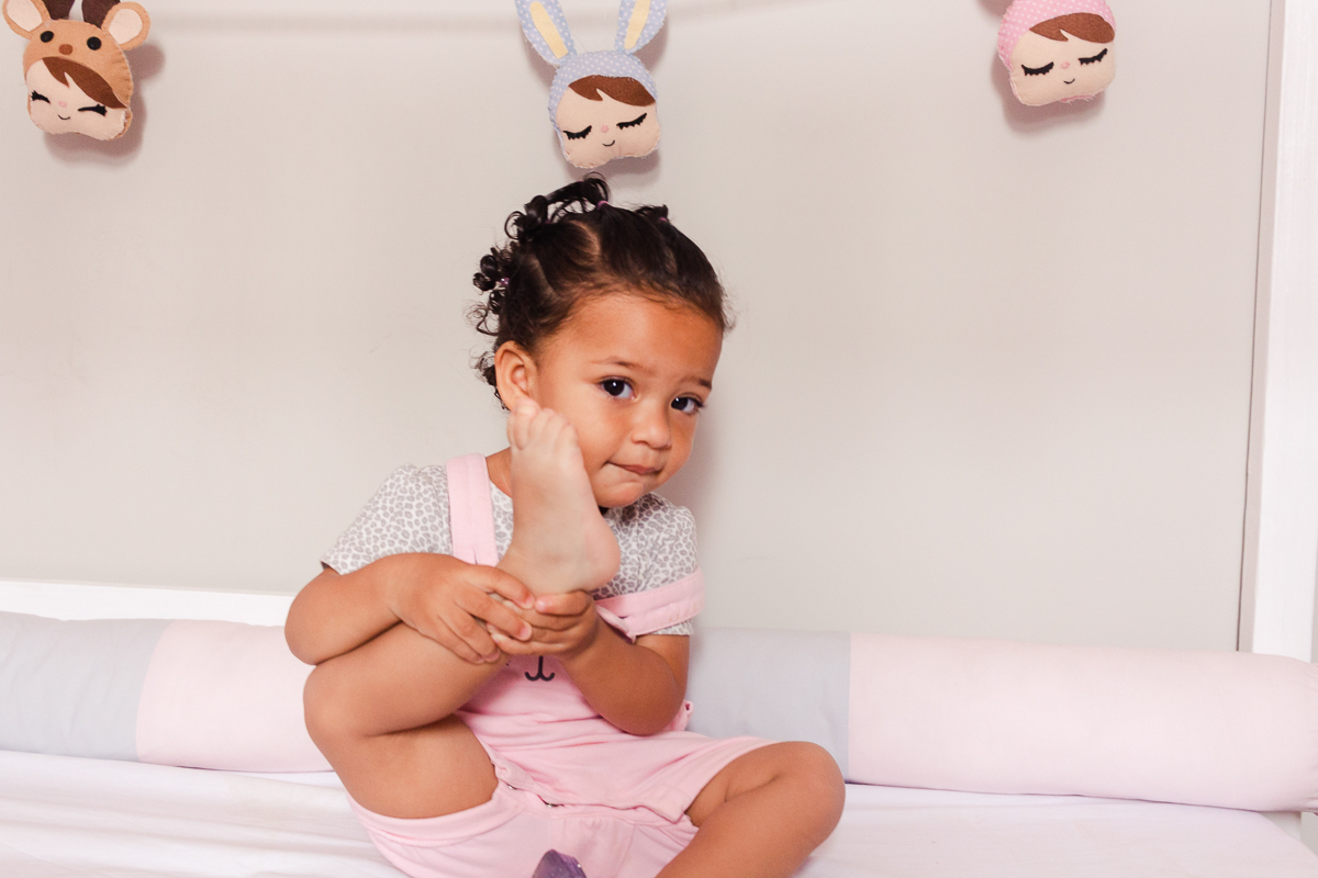 foto infantil de criança deitada dentro do foto de menina dentro do berço segurando o pé descalço, no ensaio de família em casa na zona sul da cidade de Sp