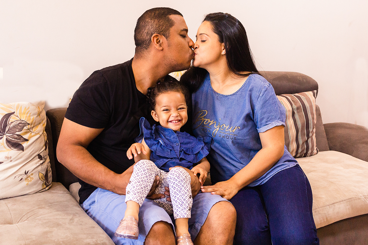 foto de pai e mãe se beijando com filha no colo na sala de casa, no ensaio lifestyle de família na zona sul de SP