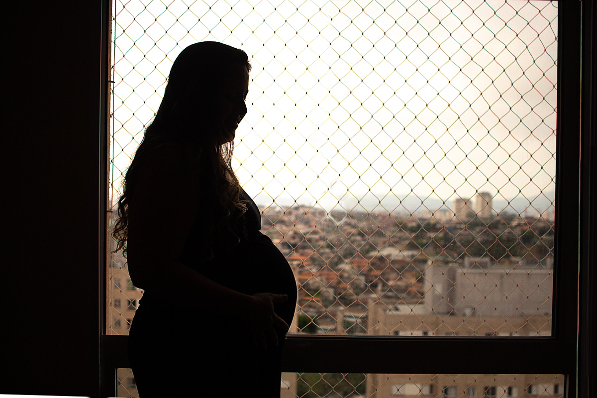 foto de silhueta de mamãe gravida com pose em frente a janela do apartamento com a mão na barriga no ensaio lifestyle de gestante na cidade de Sp 
