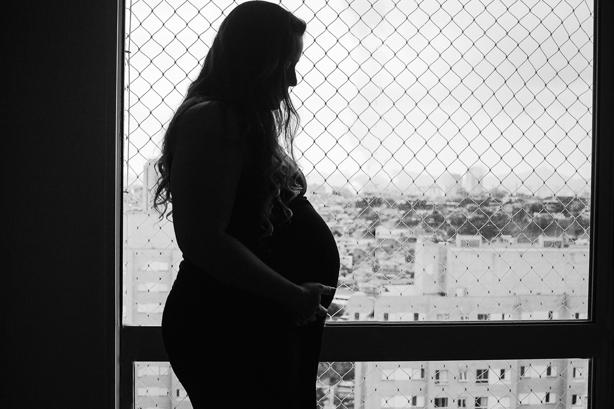 foto de silhueta de mamãe gravida com pose em frente a janela do apartamento com a mão na barriga no ensaio lifestyle de gestante na cidade de Sp 