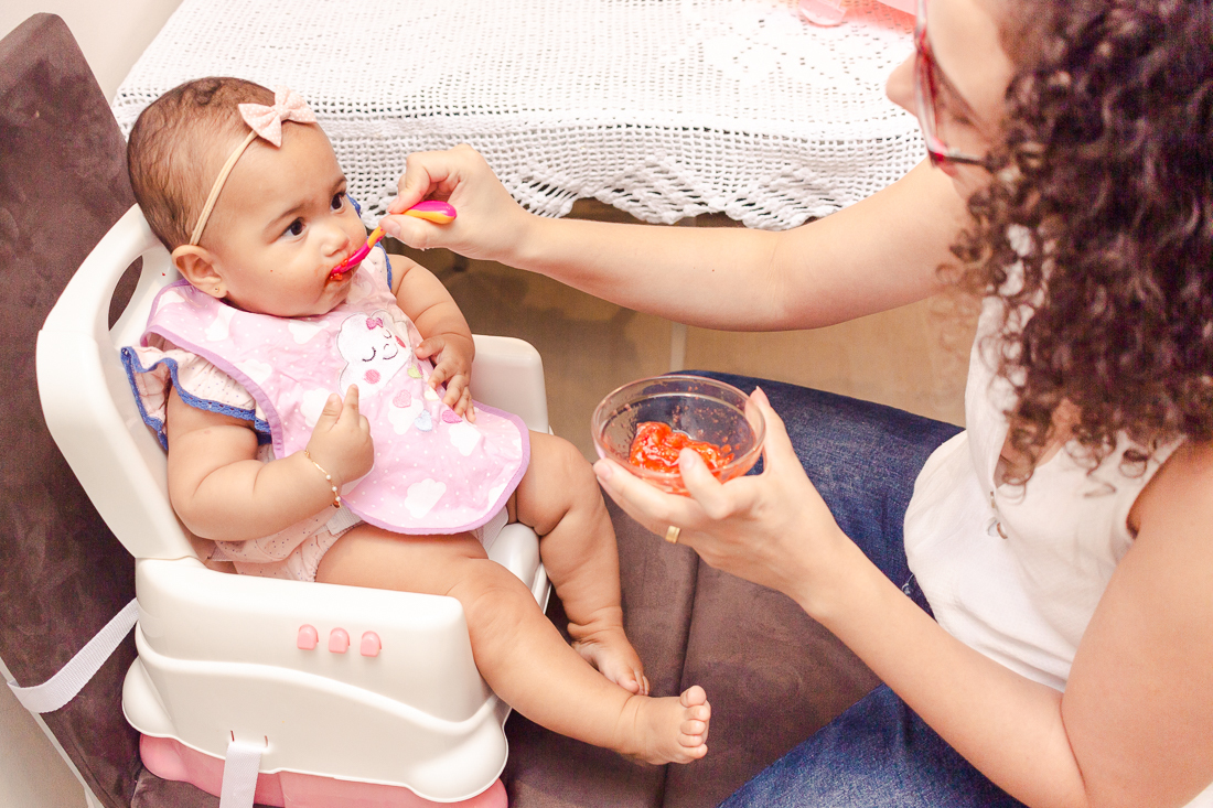 Foto da mãe dando comida para a filha na cadeirinha no ensaio na cidade de São Paulo