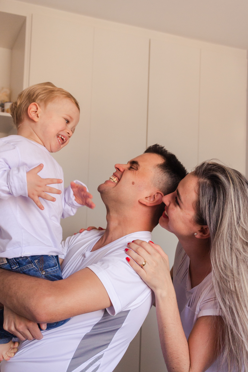 foto de criança no colo dos pais sorrindo e brincando dentro do quarto no ensaio de família em casa na cidade de Sp