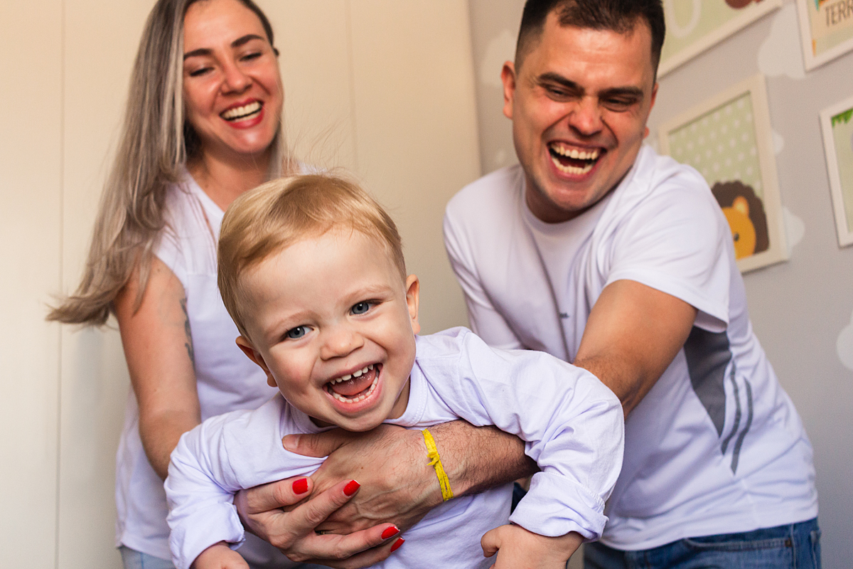 foto de criança no colo dos pais sorrindo e brincando dentro do quarto no ensaio de família em casa na cidade de Sp