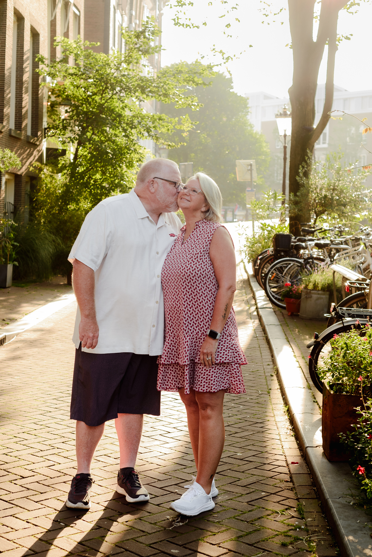 A sweet, candid moment of the couple sharing a kiss on a sunlit street in Amsterdam, with bicycles and flowers around them.