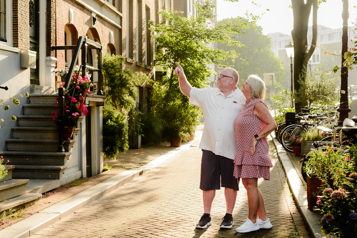 The couple standing on a sunlit street in Amsterdam, with the man pointing out a landmark as they share a moment together.