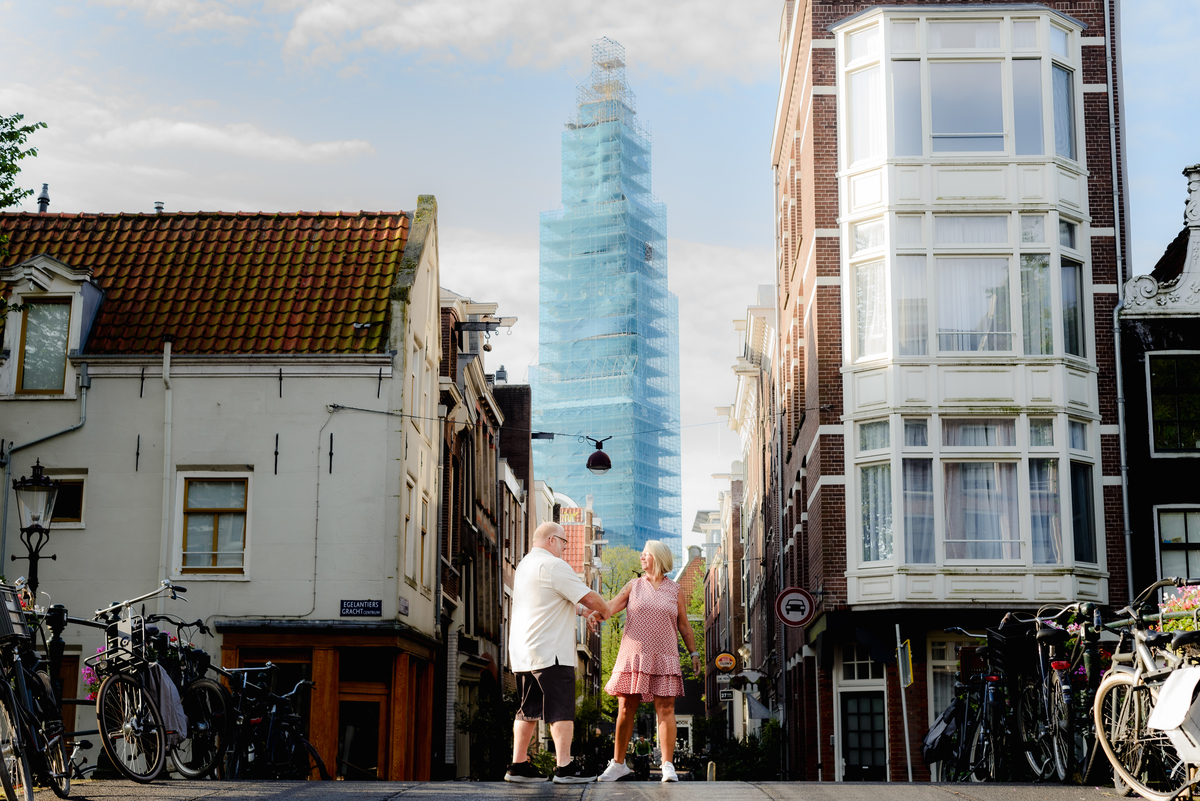 Couple standing in a quaint Amsterdam street with a towering modern building in the background, embracing and smiling.
