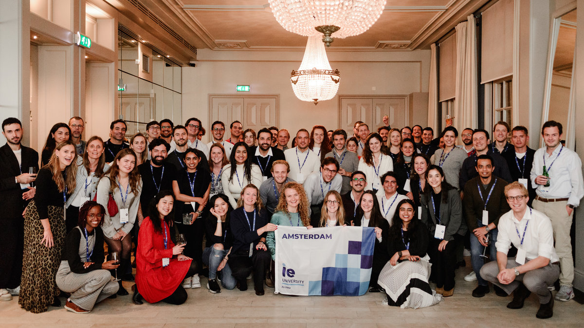 IE University-branded backdrop with alumni networking in front, capturing the professional setting of the Amsterdam event.