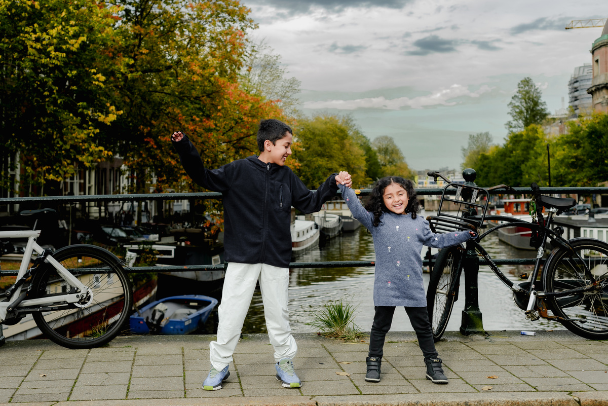 Two young kids walking hand-in-hand along an Amsterdam canal, with their family nearby, creating a playful and relaxed moment during the photoshoot.