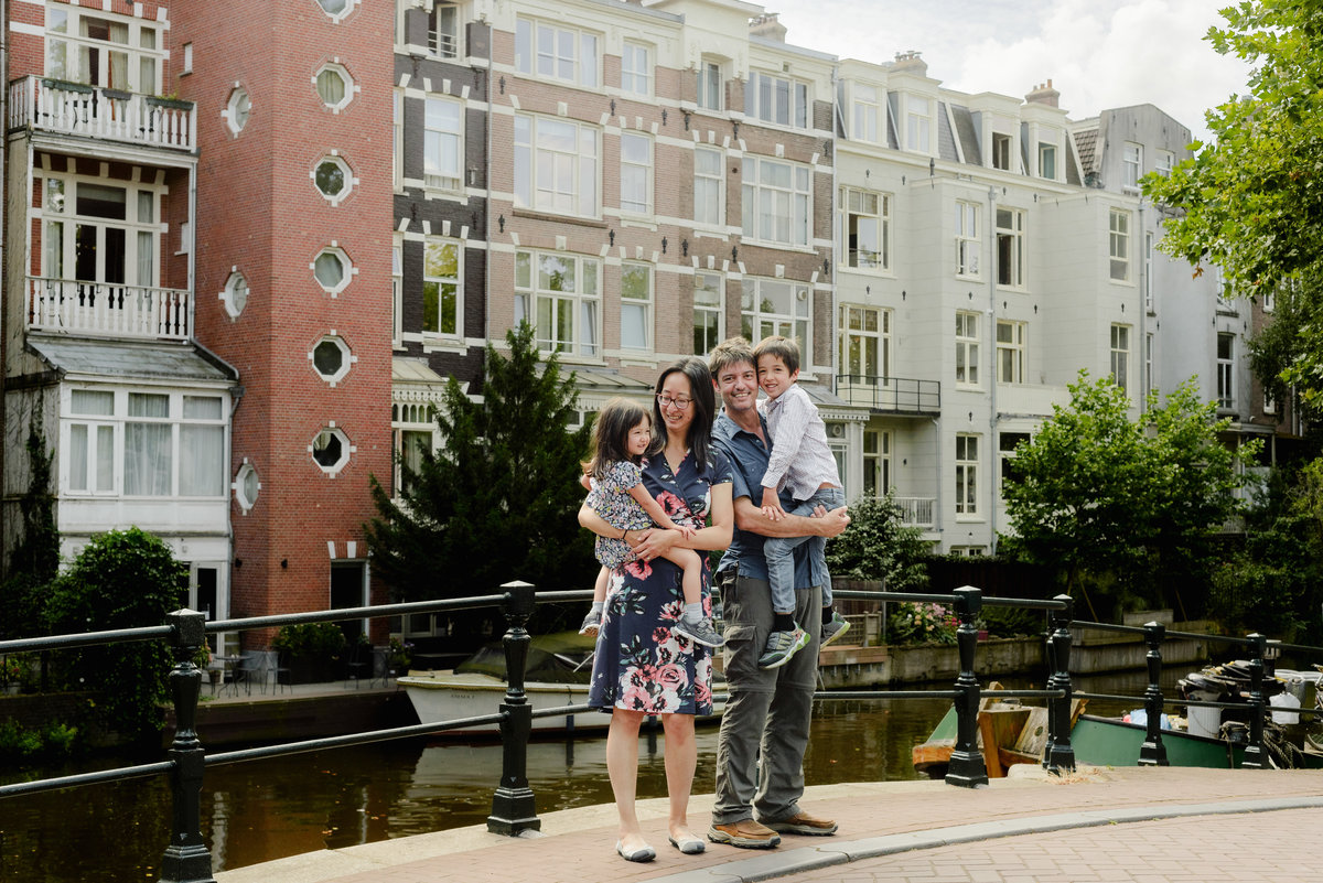 A family of four posing on a charming Amsterdam bridge with colorful houseboats in the background.