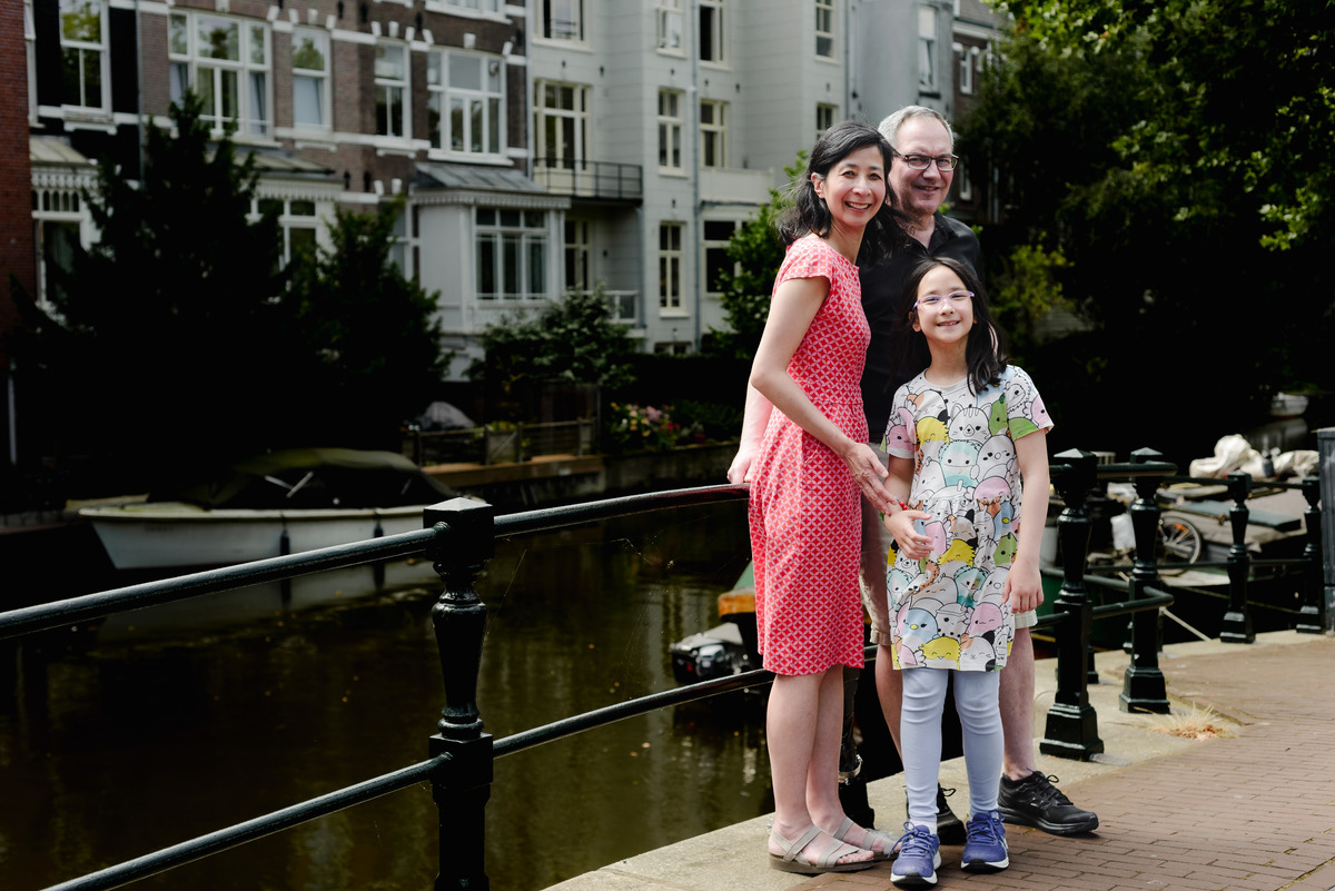 A family of three posing on a charming Amsterdam bridge with colorful houseboats in the background.