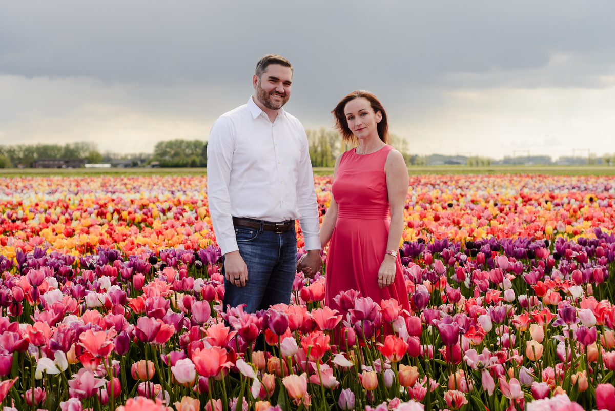 A couple stands hand in hand in a colorful tulip field under a cloudy sky, dressed in elegant attire—him in a white shirt and jeans, her in a flowing red dress.