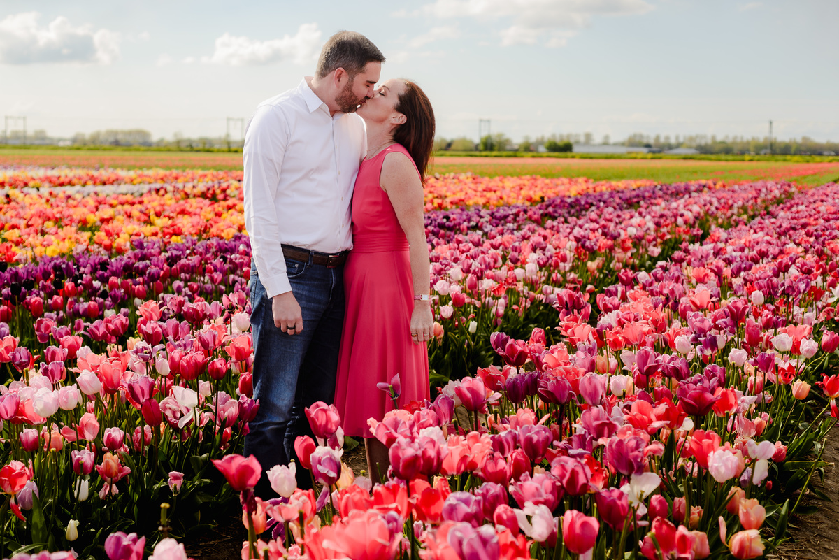 A man lovingly embraces a woman from behind as they stand in the middle of vibrant tulip rows, with red, yellow, and white flowers stretching into the horizon.