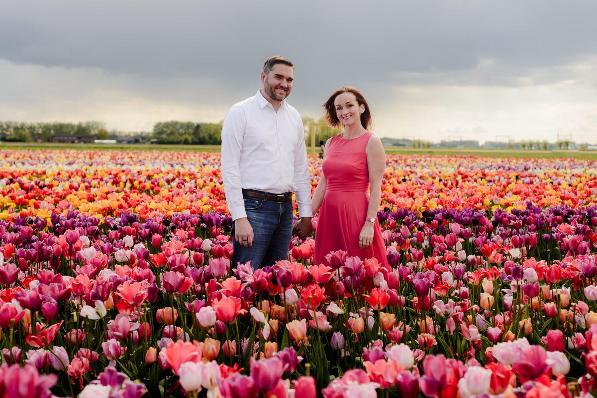 A romantic moment as a couple shares a gentle kiss while standing in a vast field of multicolored tulips under a partly cloudy sky.