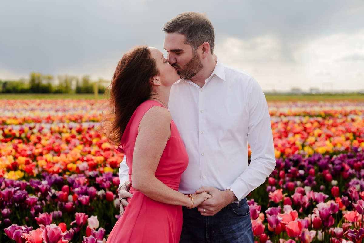 A woman in a red dress leans up to kiss her partner in a white shirt, framed by endless rows of bright tulips in full bloom.