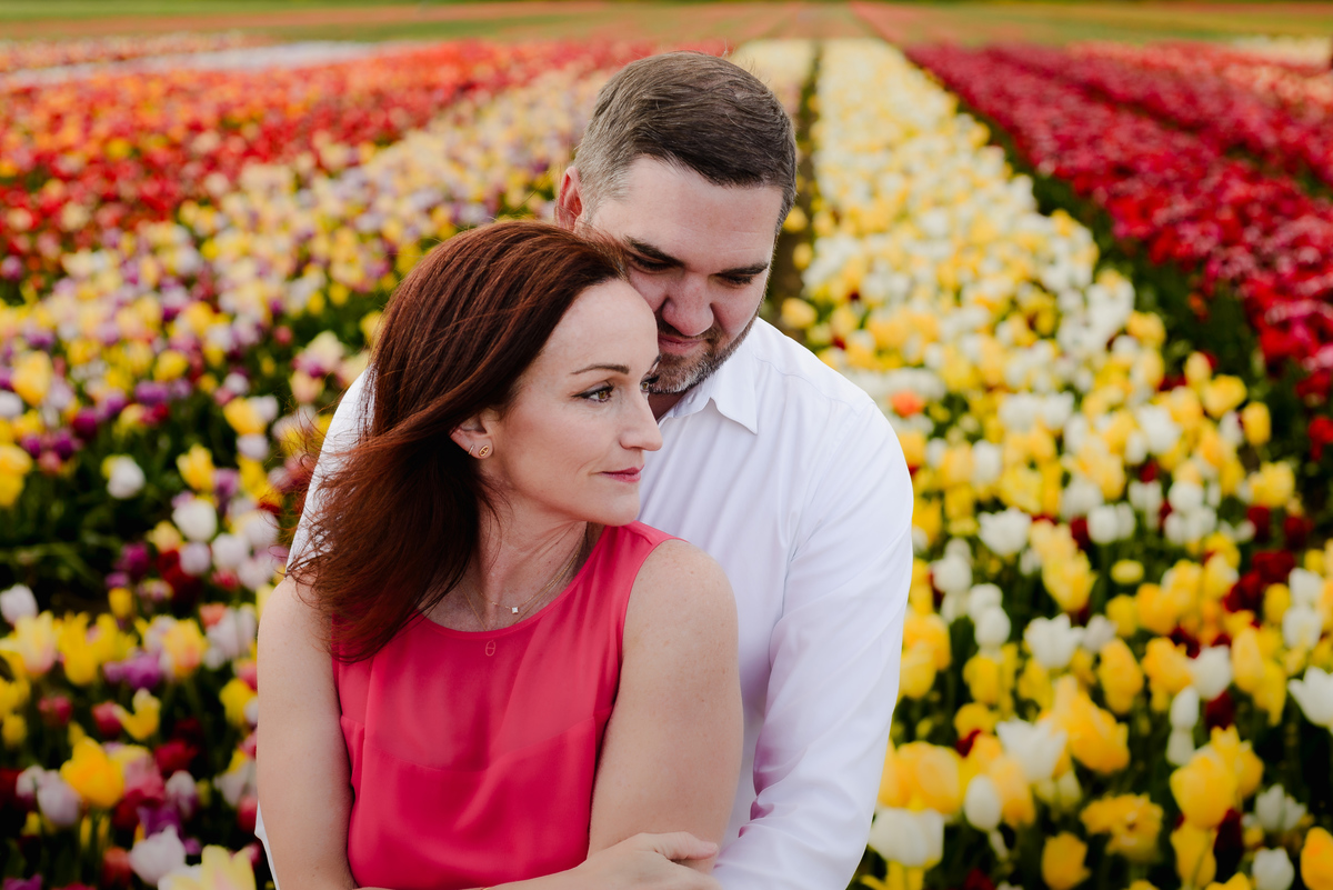 A tender embrace between a couple in a tulip field, with the woman looking into the camera as her partner holds her close against a colorful floral backdrop.