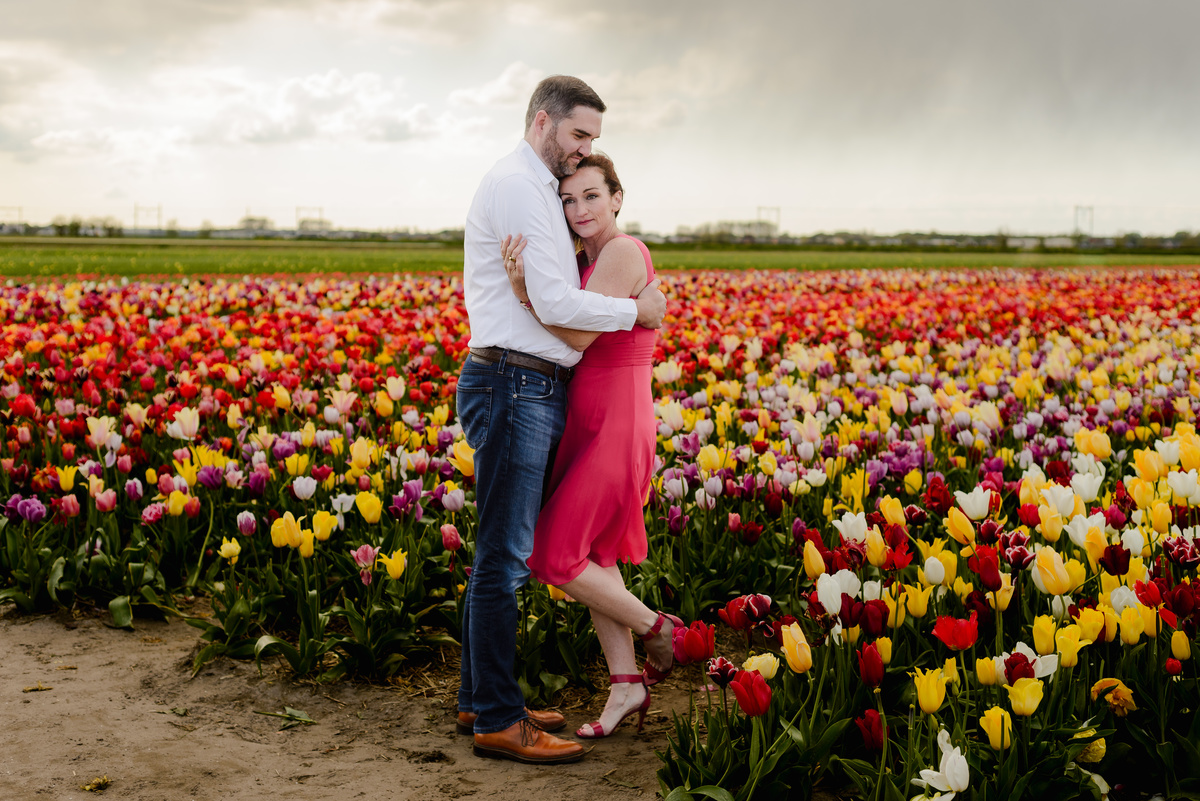 A couple walks hand in hand through a vibrant tulip field, the woman in a flowing red dress leading the way as they stroll among rows of red, yellow, and purple flowers.