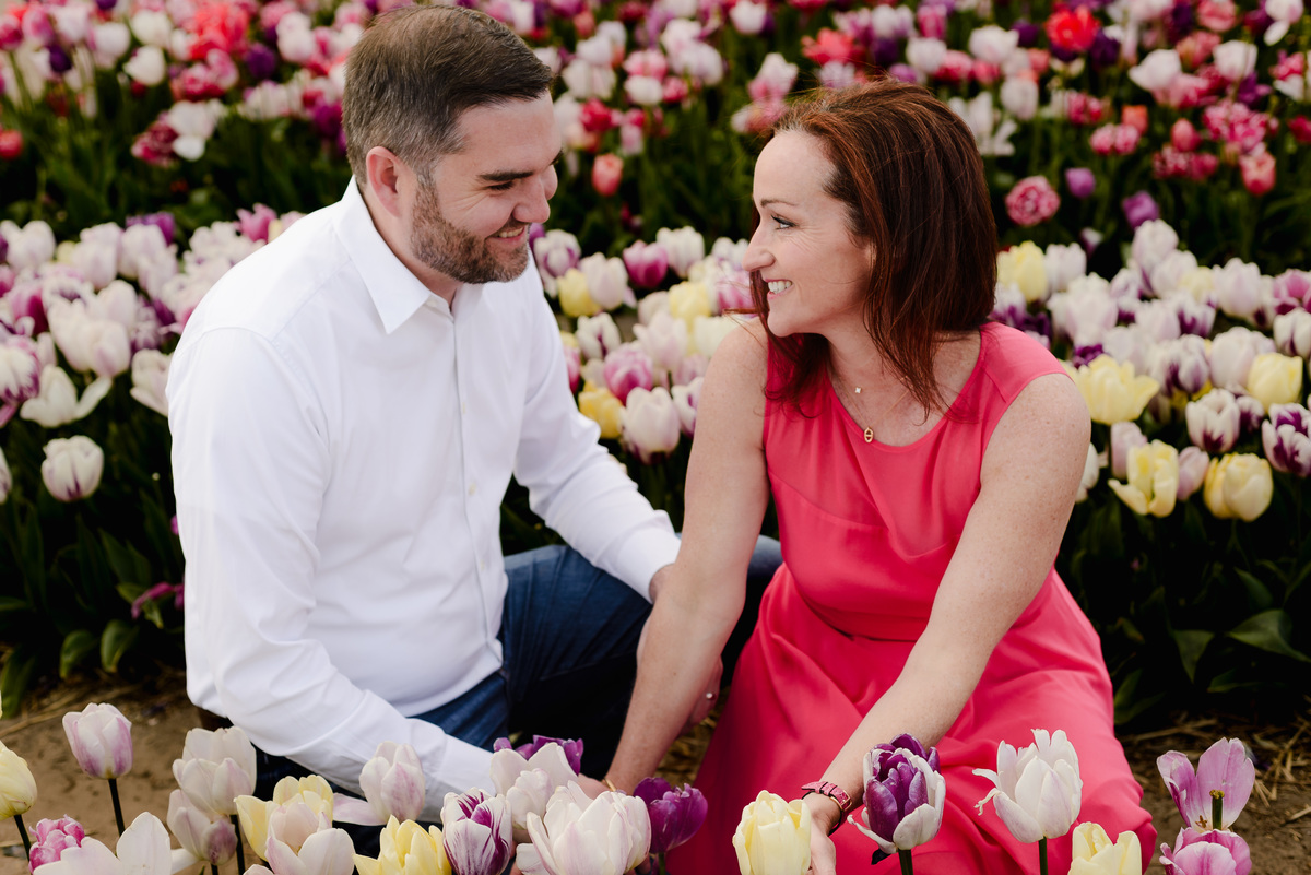 A smiling couple sits among tulip rows, facing each other with hands intertwined, surrounded by purple, yellow, and white flowers in full bloom.