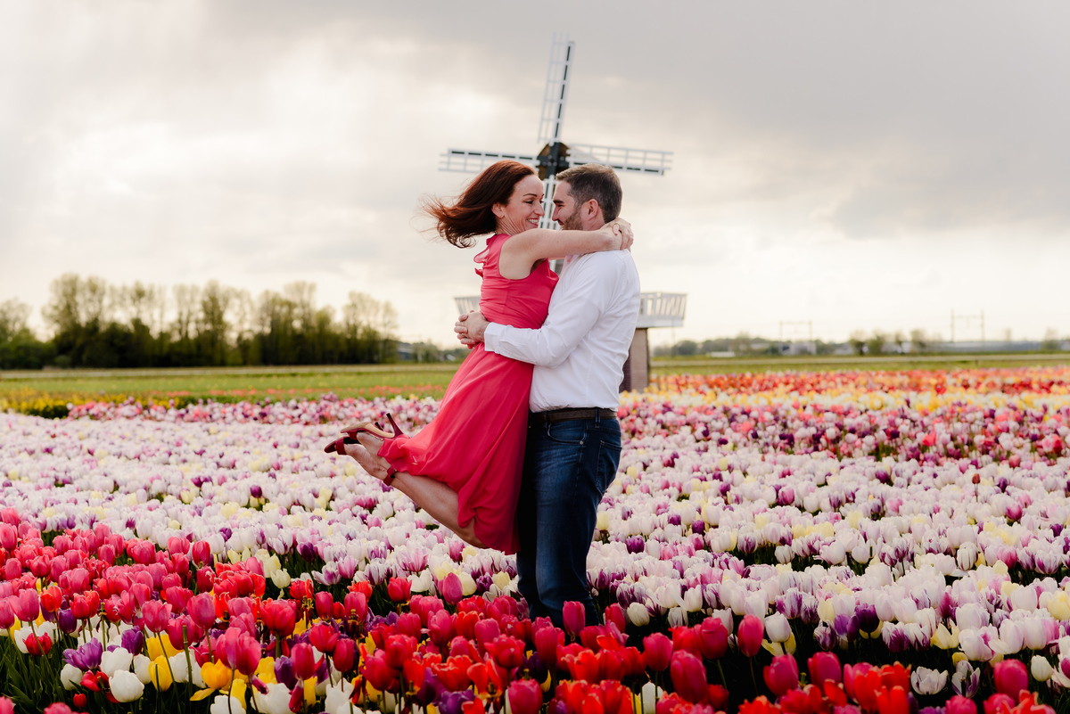 A man lifts his partner into the air as they embrace in a tulip field, with a traditional Dutch windmill in the background under a partly cloudy sky.