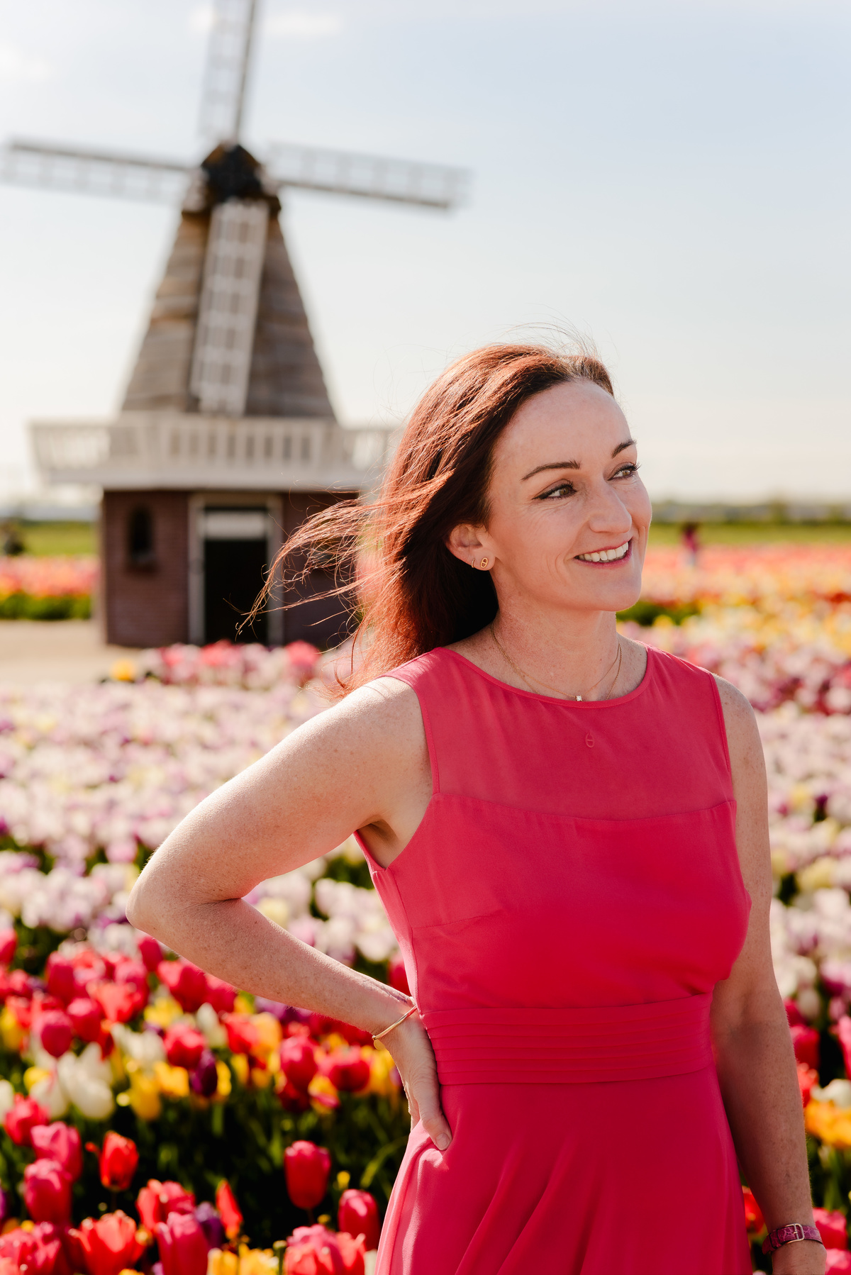 A woman in a red dress smiles warmly, standing in front of a tulip field with a windmill behind her, her hair gently blowing in the breeze.