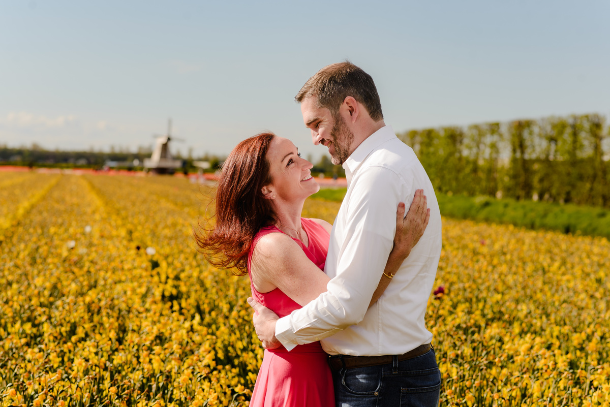 A loving embrace between a couple in the middle of a golden daffodil field, with soft sunlight highlighting their expressions and a windmill in the background.