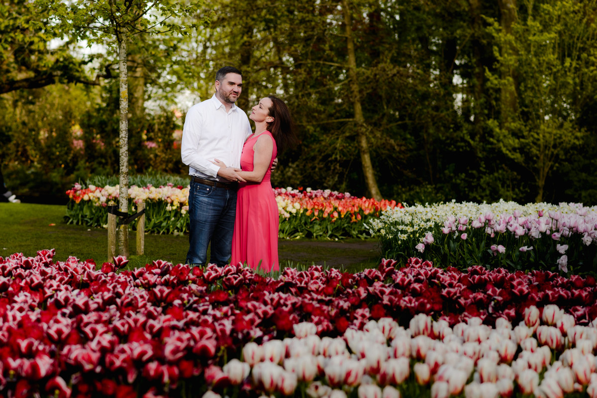 A couple stands in a lush garden, surrounded by vibrant tulips in shades of red, pink, and white, with tall green trees in the background.
