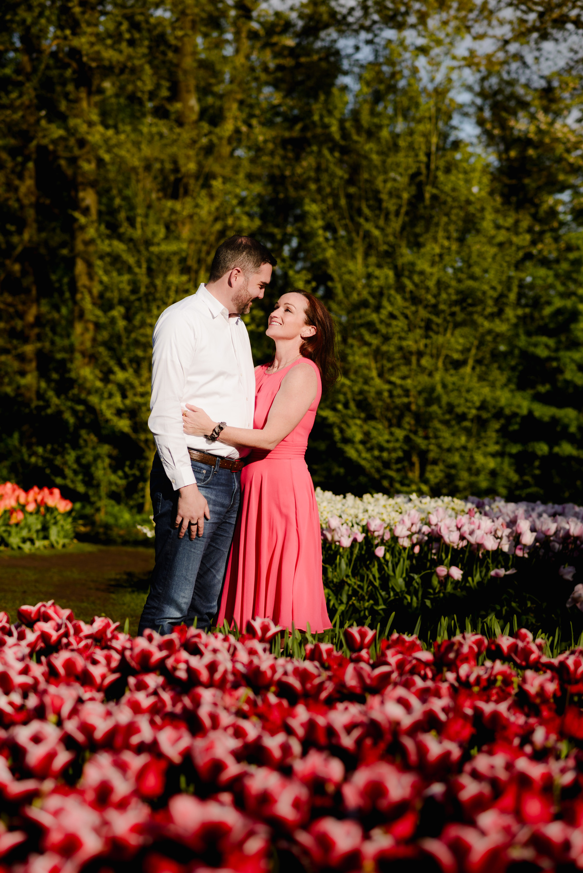 A loving moment between a couple in a flower garden, gazing into each other’s eyes while surrounded by deep red and white tulips.